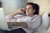Бизнесмен, использующий мобильный телефон и ноутбук в постели — стоковое фото