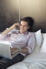 Людина використовує ноутбук комп'ютер і мобільний телефон в ліжку — стокове фото