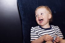 Retrato de risa niño sosteniendo chupete sentado en el sofá - foto de stock