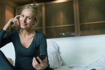 Matura seduta sul letto con smartphone in mano, sorridente, ritratto — Foto stock