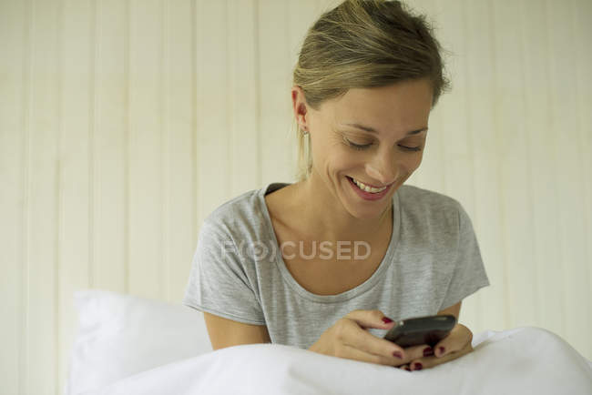 Lächelnde Frau sitzt im Bett und schreibt SMS auf dem Smartphone — Stockfoto