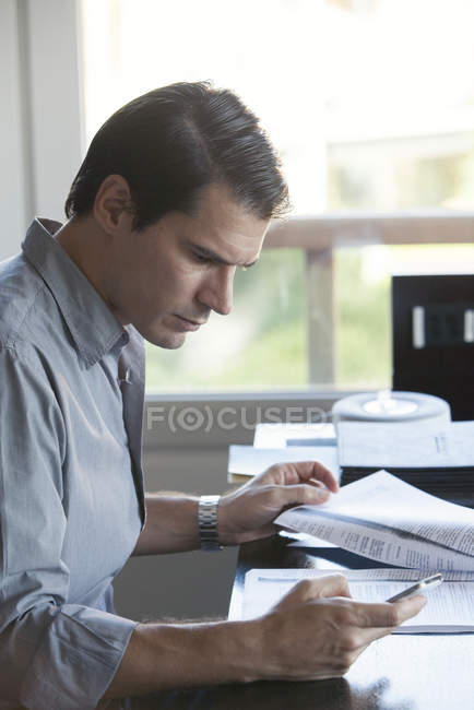 Homem olhando para o smartphone enquanto lê o documento — Fotografia de Stock