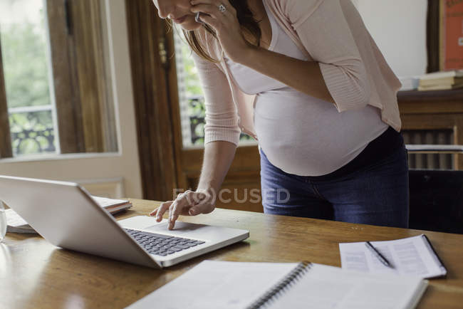 Беременная женщина пользуется ноутбуком и делает телефонный звонок — стоковое фото