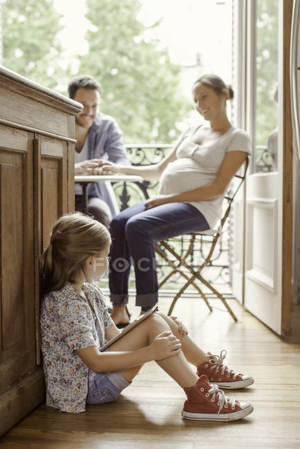 Chica sentada en el suelo con la tableta digital como los padres de chat en segundo plano - foto de stock