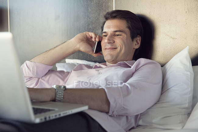 Empresário usando telefone celular e computador portátil na cama — Fotografia de Stock