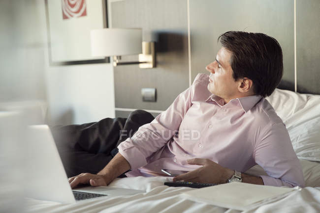Empresário deitado na cama do hotel com telefone celular e computador portátil, olhando para longe no pensamento — Fotografia de Stock