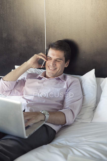 Homem usando computador portátil e telefone celular na cama — Fotografia de Stock