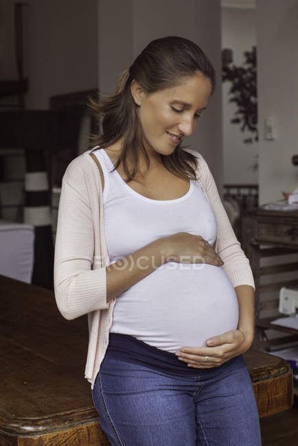 Retrato de la mujer embarazada cogida de la mano en el estómago en casa - foto de stock
