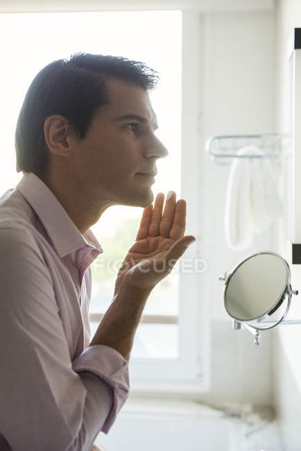 Человек, прикладывающий увлажняющий крем к лицу, глядя в зеркало — стоковое фото
