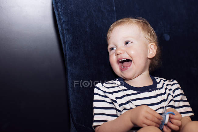 Ritratto di ridere bambino che tiene ciuccio seduto sul divano — Foto stock