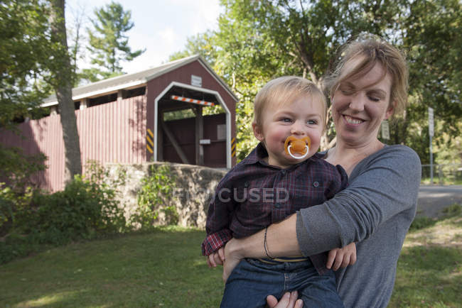 Portrait of Mother holding toddler son in front of covered bridge — Stock Photo