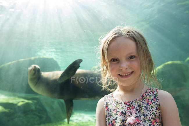 Portrait of smiling little girl at aquarium with seal swimming on the background — Stock Photo