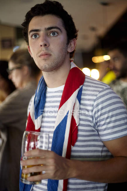 Tifoso di calcio britannico che guarda la partita nel bar con bandiera avvolta intorno al collo e vetro di birra — Foto stock