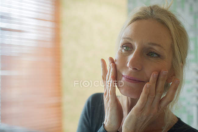 Зріла жінка дивиться на своє відображення в дзеркалі з руками на щоках — стокове фото