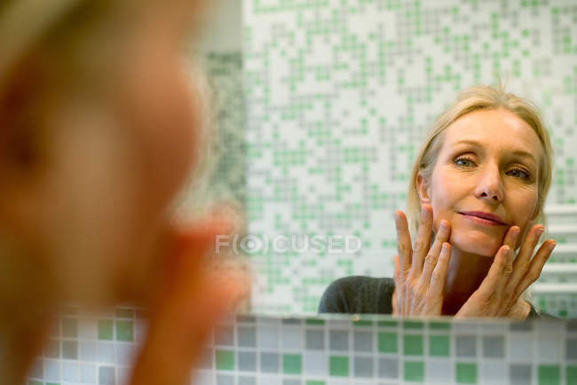 Donna matura scrutando il suo viso nello specchio del bagno — Foto stock
