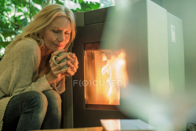 Mulher madura relaxante por lareira com bebida quente — Fotografia de Stock