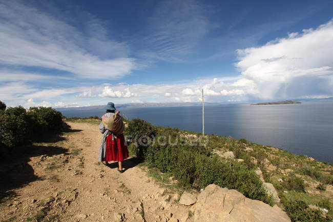 Rückansicht einer Frau in traditioneller bolivianischer Kleidung, die an einem sonnigen Tag an einem Felsen über das Meer wandert — Stockfoto