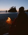 Retrato trasero de una mujer sentada en la playa de arena cerca de una hoguera al atardecer. Segunda playa, Península Olímpica, La Push, Washington - foto de stock