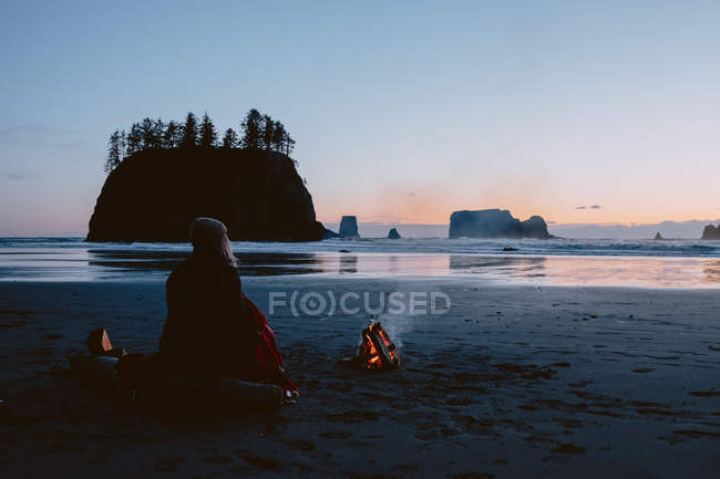 Retrato trasero de una mujer sentada en la playa de arena cerca de una hoguera al atardecer. Segunda playa, Península Olímpica, La Push, Washington - foto de stock