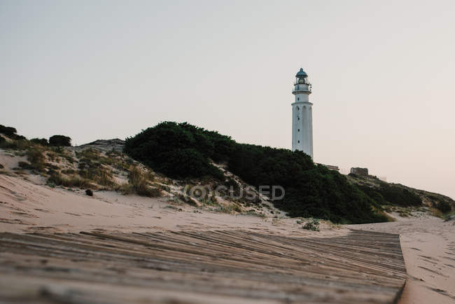 Bâtiment phare sur la plage de sable vue au coucher du soleil et passerelle en bois au premier plan — Photo de stock