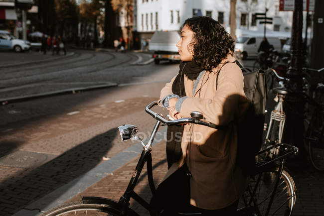Vista lateral de mujer joven con bicicleta a la luz del sol en la calle de la ciudad - foto de stock
