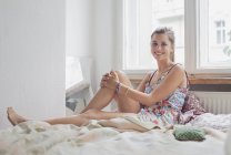 Portrait of smiling young woman sitting on bed against window at home — Stock Photo