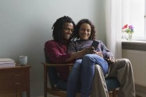 Couple souriant multi-ethnique utilisant un téléphone intelligent tout en étant assis sur un fauteuil à la maison — Photo de stock