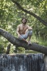 Homme torse nu réfléchi assis sur une branche au-dessus d'une cascade dans la forêt — Photo de stock