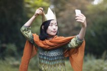 Retrato de mujer con corona de papel tomando selfie con smartphone en el parque - foto de stock