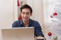 Sorridente giovane uomo seduto vicino all'albero di Natale artificiale e computer portatile di navigazione — Foto stock