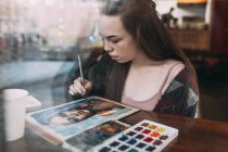 Brunette woman painting at cafe seen through window — Stock Photo