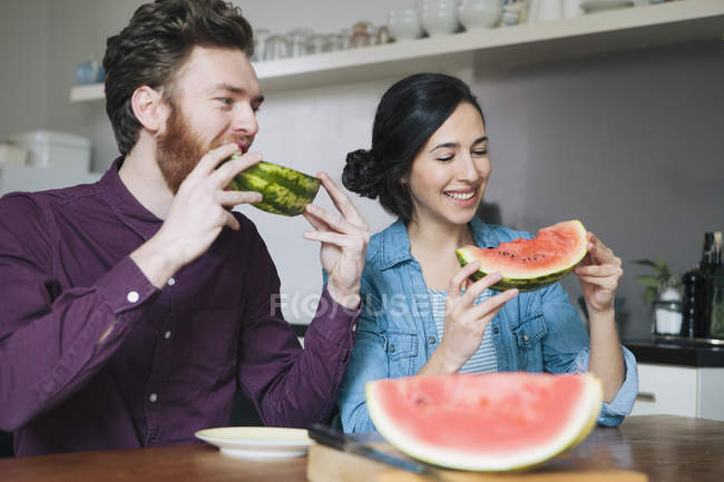 Feliz pareja joven comiendo sandía en la mesa de la cocina - foto de stock