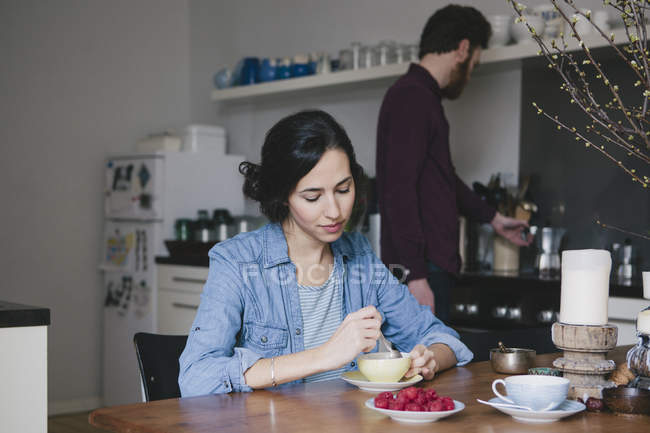 Young woman stirring coffee at kitchen table with man on background — Stock Photo