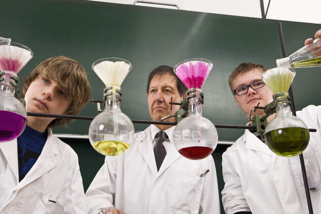 Un profesor que supervisa a dos estudiantes que realizan un experimento de química - foto de stock