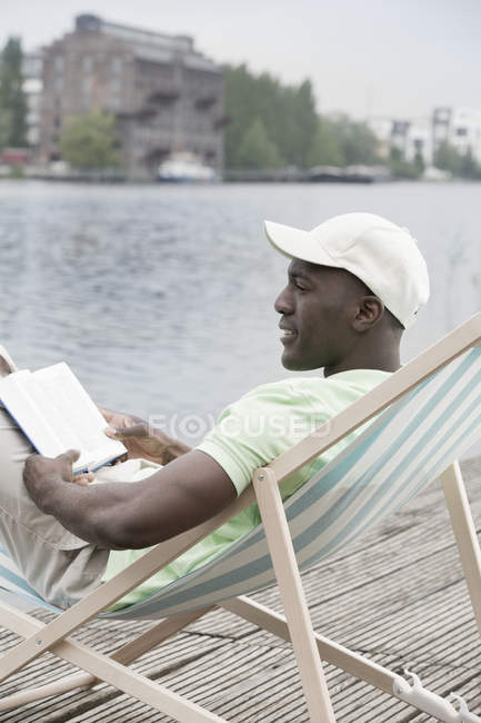 Mann entspannt mit Buch auf Liegestuhl am See — Stockfoto