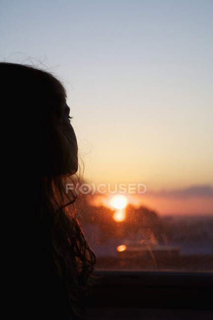 Uma rapariga a olhar pela janela ao pôr do sol — Fotografia de Stock