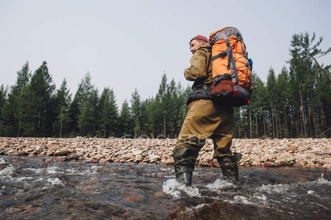 Low angle view of mid adult man with backpack standing in river at forest — Stock Photo