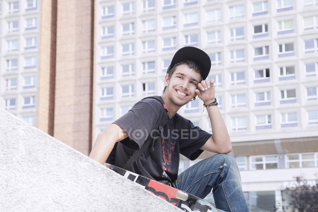 Happy young man with skateboard sitting against building — Stock Photo