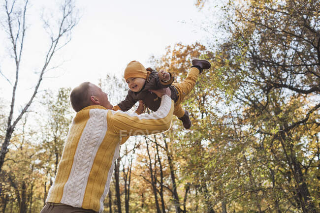Vista de ángulo bajo del alegre hombre que levanta al niño en el parque durante el otoño - foto de stock