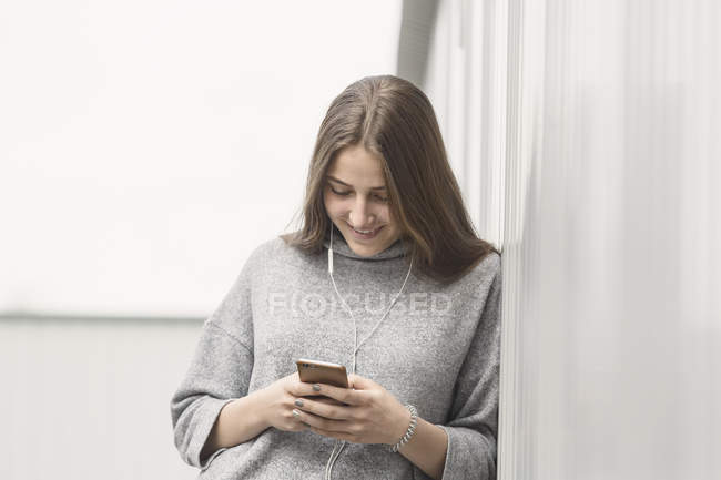 Улыбающаяся девушка-подросток с помощью мобильного телефона, опираясь на стену — стоковое фото