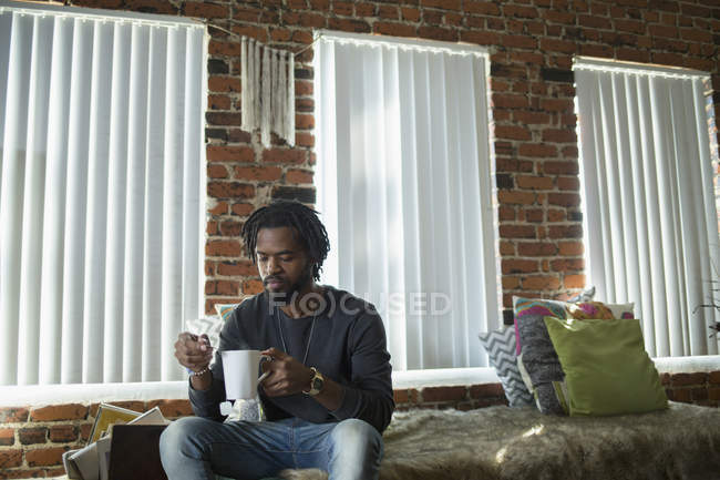 Uomo seduto sul letto con tazza di caffè contro le finestre a casa — Foto stock