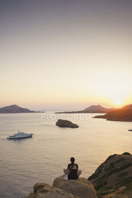 Rear view of woman sitting on rock formation while looking at sea against sky during sunset — Stock Photo