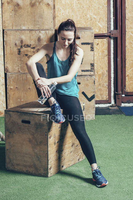 Полная длина женщины-спортсмена, держащей бутылку воды, сидя на деревянной коробке в спортзале — стоковое фото