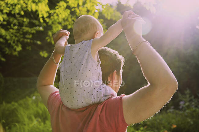 Vista trasera del padre cargando al niño en hombros en el patio durante el verano - foto de stock