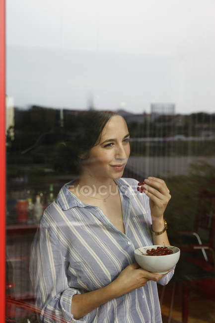 Mujer joven pensativa comiendo grosellas rojas mientras mira a través de la ventana - foto de stock