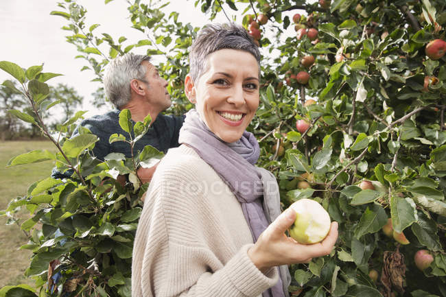 Retrato de mujer feliz comiendo manzana en huerto con el hombre en el fondo - foto de stock