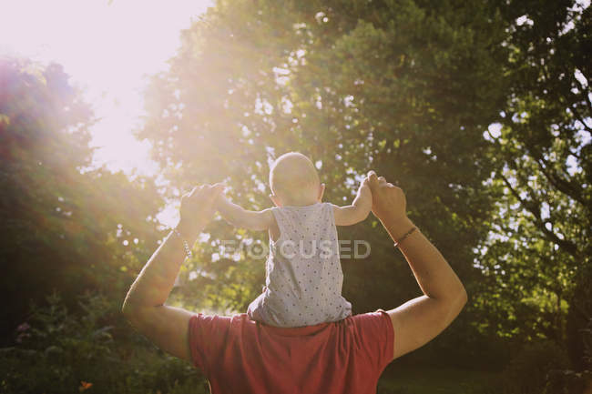 Rear view of father carrying baby boy on shoulders at yard during sunny day — Stock Photo