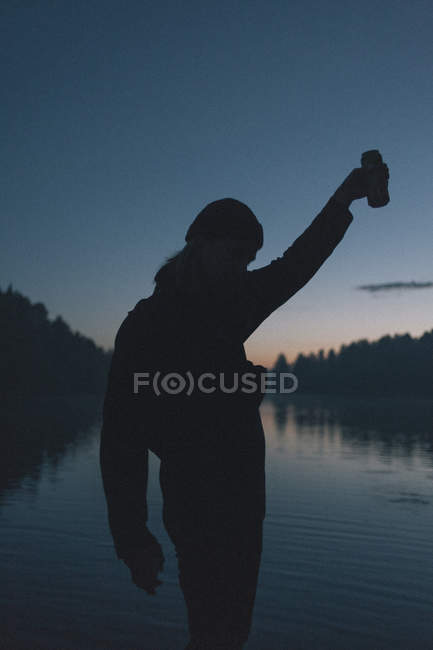 Silueta de mujer sosteniendo la bebida puede contra el lago al atardecer - foto de stock