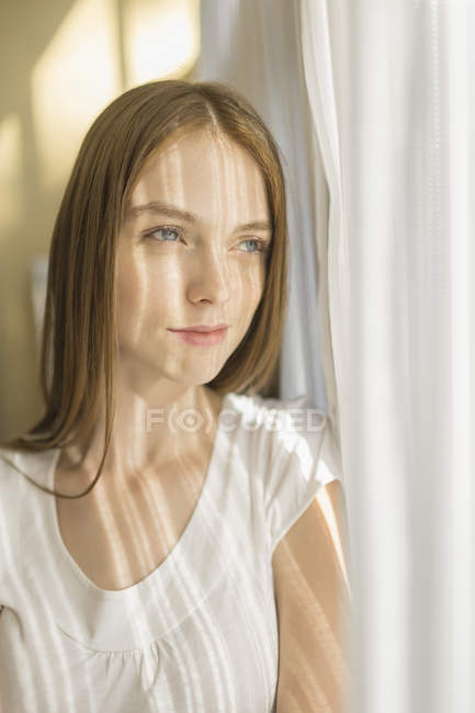 Mujer sonriente reflexiva de pie junto a las cortinas en casa en el día soleado - foto de stock