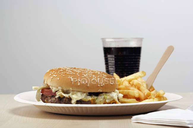 Burger and french fries on paper plate next glass of cola on table — Stock Photo